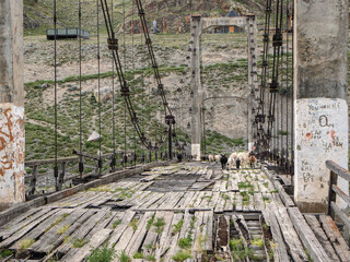 A weathered wooden suspension bridge in a mountain valley is crossed by a herd of goats.