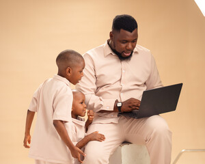 African dad uses a laptop while his two sons watch attentively. Dressed in matching beige outfits,...