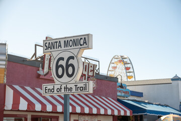 Iconic Route 66 &lsquo;End of the Trail&rsquo; sign at Santa Monica Pier with Ferris wheel and striped canopies in the background