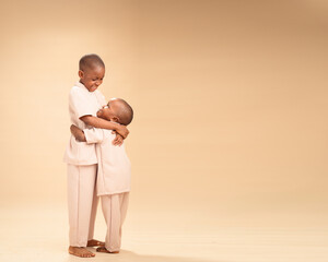 Two young African boys in matching cream outfits share a joyful hug in a studio setting. The older...