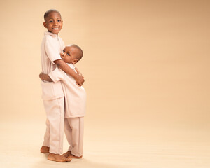 Two young African brothers wearing matching beige outfits hug and smile warmly while standing...