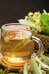 Tasty linden tea in cup, leaves and flowers on wooden table against dark background, closeup