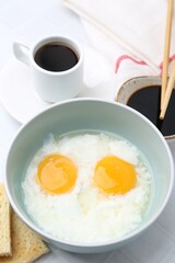 Traditional asian breakfast with half-boiled eggs in bowl, soy sauce, bread, chopsticks and coffee on white table, above view