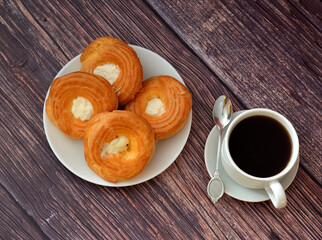 A cup of black coffee on a saucer with a spoon and a plate with four custard cakes with protein cream on a wooden table.