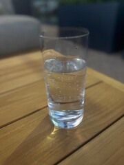 A clear glass of carbonated water resting on a sunlit wooden surface with natural shadows and soft-focus background. Refreshing and minimal stock photo.