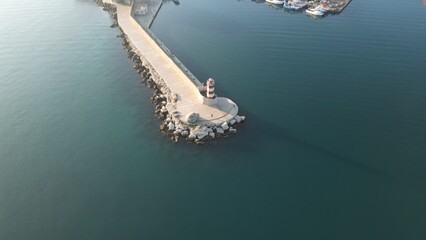 Coastal Beacon: An aerial perspective unveils a sturdy stone pier reaching into tranquil azure waters, culminating in a classic lighthouse that guides ships through the open ocean. Monopoli, Puglia
