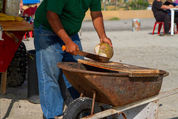 Close up of a man chopping a coconut with a macheti on a wheelbarrow on a beach