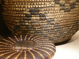 Dominating the frame, a large, intricately woven basket contrasts with a smaller decorated plate on a table, highlighting craftsmanship and design