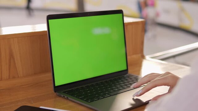Person is typing on a laptop with a green screen. The laptop is on a wooden table