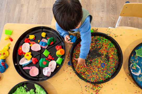 Overhead view of a young child or toddler participating in sensory play with colorful rice, playdough on a table.Concept of fine motor skills, hands-on learning, childcare center, or day care activity