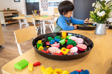 Colorful playdough in an early learning preschool classroom or activity table features various tools and shapes for sensory exploration with a young child or toddler playing in the background.