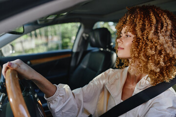 Curly woman driving a car with focused expression, wearing a seatbelt in a modern vehicle interior, natural daylight, spring season outside the window.