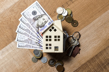 US Dollar Bills Fanned Out Behind Small House Model, Surrounded By Various Coins On Wooden Table