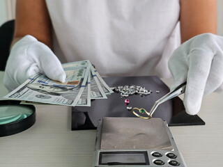 Jeweler weighing a ring with an emerald surrounded by cash and gemstones at a worktable