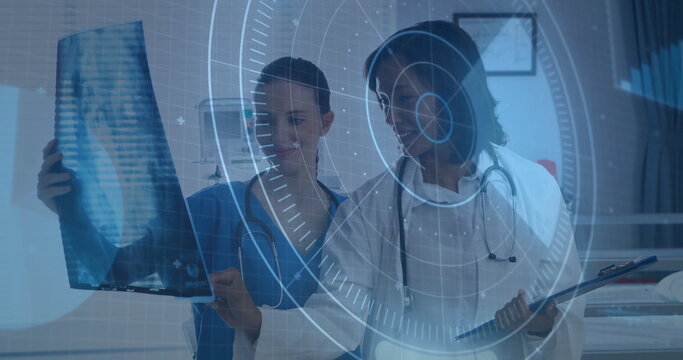 Two medics in scrubs holding X-ray film and reviewing clipboard in exam room with digital overlay