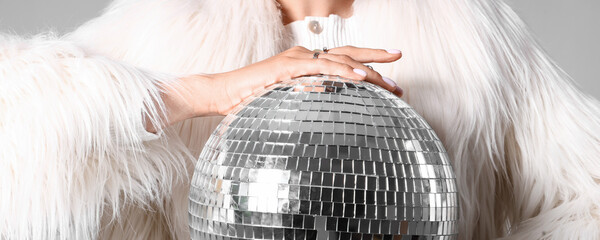 Fashionable young woman in fur coat with disco ball on grey background