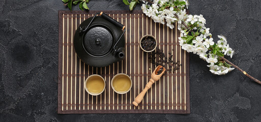 Teapot with cups, wooden spoon of dry tea and flowers on black grunge background