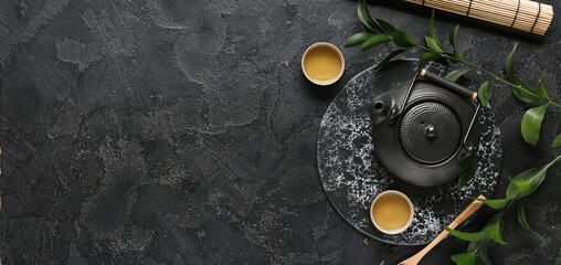 Teapot, cups of hot tea and spoon with dry leaves on black background