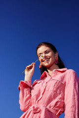 Smiling female model with flower against a blue sky, wearing a pink dress, showcasing joy and...