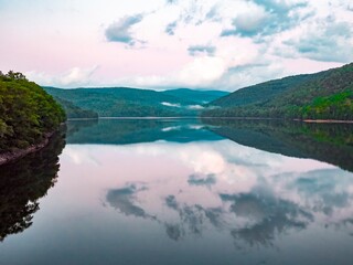 Dramatic sky over calm lake, with reflections of forest covered hills and clouds in the water, purple- late afternoon sky and mist in the distance. Pepacton Reservoir, Catskills, New York State USA.