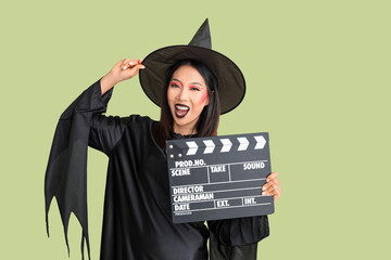Beautiful young Asian woman dressed for Halloween as witch with clapperboard on green background