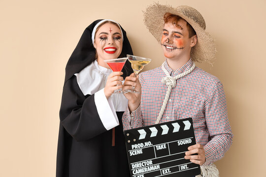 Young couple in costumes for Halloween party with clapperboard and cocktails on beige background