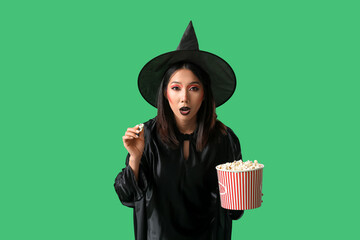 Shocked young Asian woman dressed for Halloween as witch with popcorn on green background