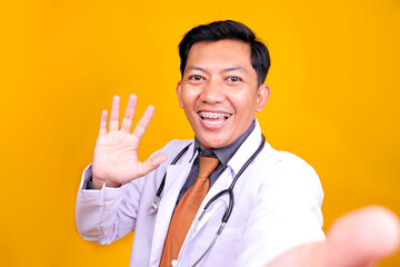 Young Asian Male Doctor In A White Coat Waves Directly At The Camera Over Yellow Background
