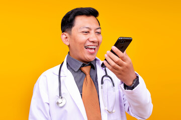 Cheerful Male Doctor In A White Coat Laughs While Looking At His Phone Isolated On Yellow Background