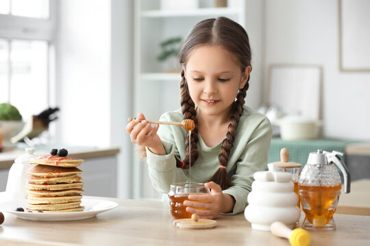 Cute little girl with honey and pancakes at table in kitchen