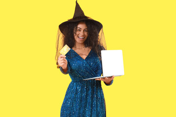 Young African-American woman dressed for Halloween as witch with credit card and laptop shopping...