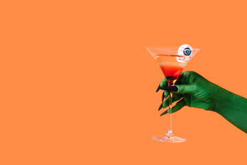 Green hand of witch with glass of cocktail on orange background. Halloween celebration