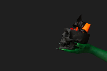 Naklejka premium Green hand of witch holding skull with candies and gift box on black background. Halloween celebration