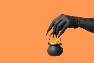 Black hand of witch with cauldron on orange background. Halloween celebration