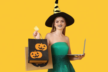 Young woman dressed for Halloween as witch with laptop and shopping bags on yellow background