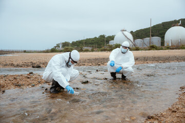 Two scientists collect water samples on a beach near industrial facilities during a cloudy day for...