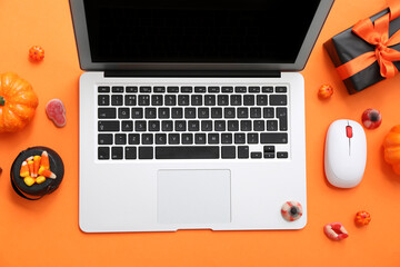 Laptop with Halloween candies and gifts on orange background
