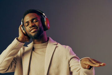 Young man enjoying music with headphones, wearing a stylish beige coat against a gradient...