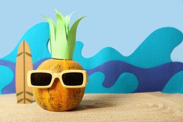 Halloween pumpkin, surfboard and sunglasses on sand against blue background