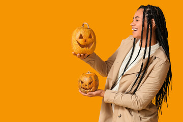 Beautiful young happy African-American woman with carved Halloween pumpkins on yellow background