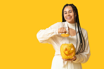 Beautiful young happy African-American woman with carved Halloween pumpkin on yellow background