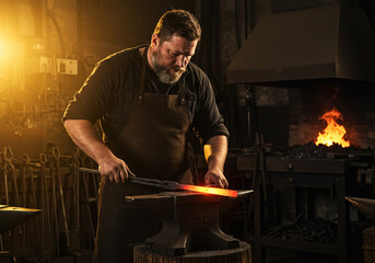 Bearded blacksmith forging red hot metal on anvil in a dark workshop with fire glow