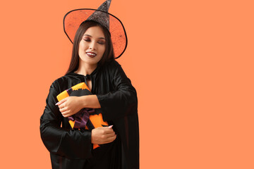 Beautiful young happy Asian woman dressed as witch for Halloween with gift box on orange background
