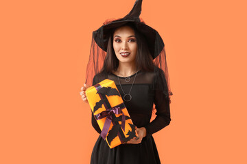 Beautiful young happy Asian woman dressed as witch for Halloween with gift box on orange background