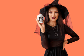 Beautiful young happy Asian woman dressed as witch for Halloween with human skull on orange...