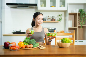 Smiling Asian woman in sporty wear eating fresh salad at table with fruits, veggies, and green juice. Promotes clean eating, wellness, and a healthy lifestyle concept.