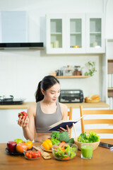 Asian woman in fitness wear uses tablet while eating healthy food at kitchen table. Holds tomatoes, writes in notebook. Clean eating and remote wellness lifestyle theme.