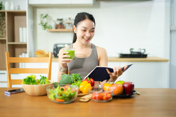 Asian woman in fitness wear uses tablet while eating healthy food at kitchen table. Holds tomatoes, writes in notebook. Clean eating and remote wellness lifestyle theme.
