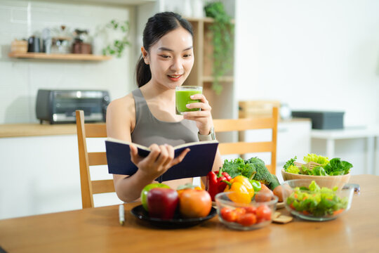 Asian woman in fitness wear uses tablet while eating healthy food at kitchen table. Holds tomatoes, writes in notebook. Clean eating and remote wellness lifestyle theme. - Powered by Adobe