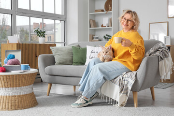 Beautiful middle-aged woman with cute cat and cup of tea sitting on sofa at home
